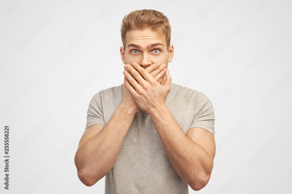 Incredulous attractive man covers mouth, doesnt want to spread rumours, shocked to hear sudden news from interlocutor, dressed in casual tshift isolated on white wall - obrazy, fototapety, plakaty 