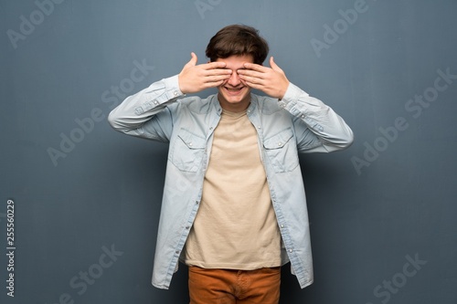 Teenager man with jean jacket over grey wall covering eyes by hands. Surprised to see what is ahead