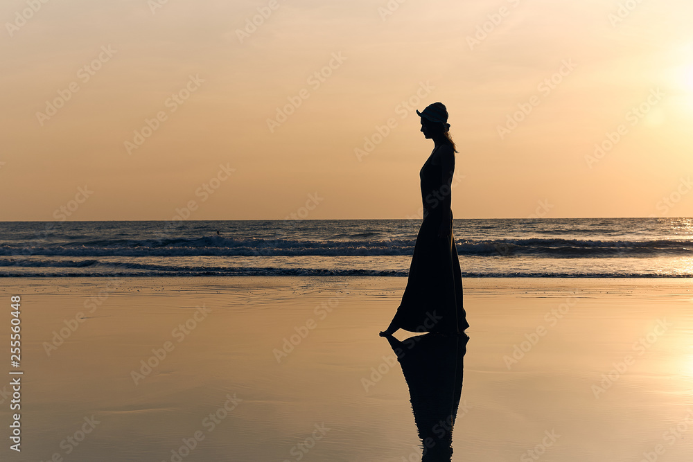 Contrast silhouette of a young slender woman against the background of a Sunny sunset, the sea and the sandy beach. Warm evening tones and reflections of the figure on the wet sand