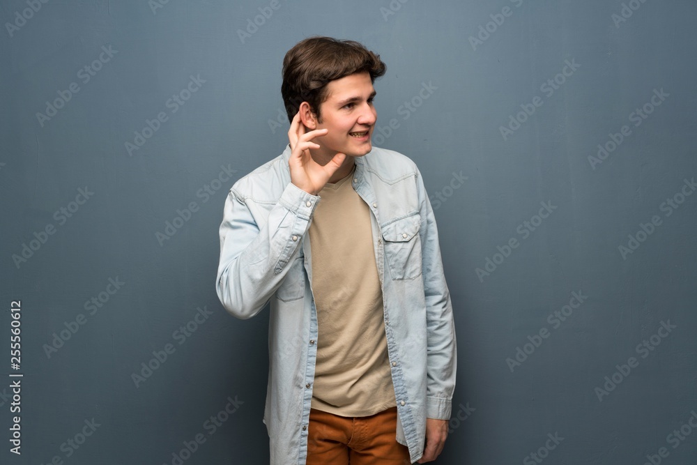 Teenager man with jean jacket over grey wall listening to something by putting hand on the ear