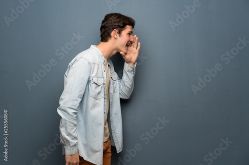 Teenager man with jean jacket over grey wall shouting with mouth wide open to the lateral