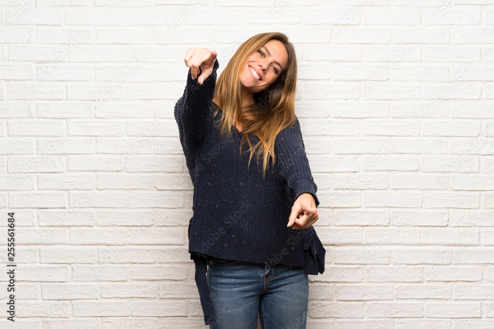 Blonde woman over brick wall points finger at you while smiling Blonde woman over brick wall points finger at you while smiling