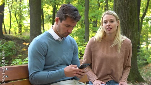 A man and a woman sit on a bench in a park, the man is absorbed with his smartphone, the woman is angry at him