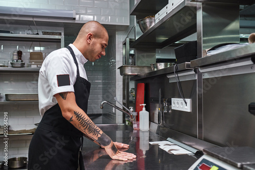 Let's start. Young bald male chef with beautiful tattoos on his arms looking at order lists on a steel table in a restaurant kitchen.