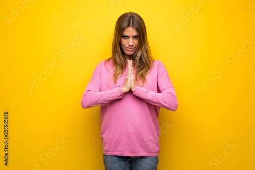 Woman with pink sweater over yellow wall keeps palm together. Person asks for something