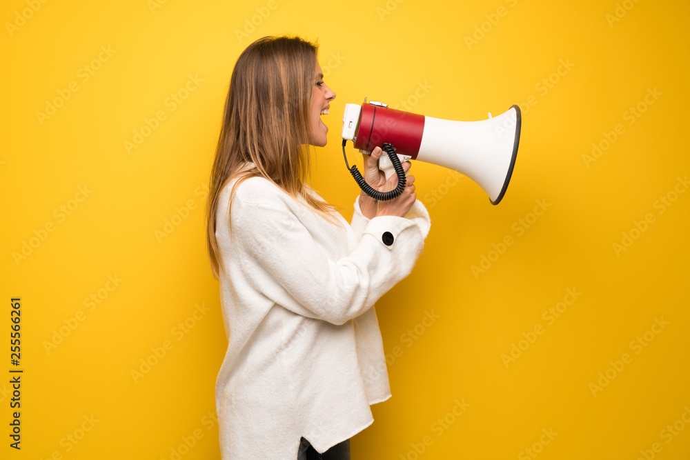 Blonde woman over yellow wall shouting through a megaphone to announce something in lateral position Blonde woman over yellow wall shouting through a megaphone to announce something in lateral position