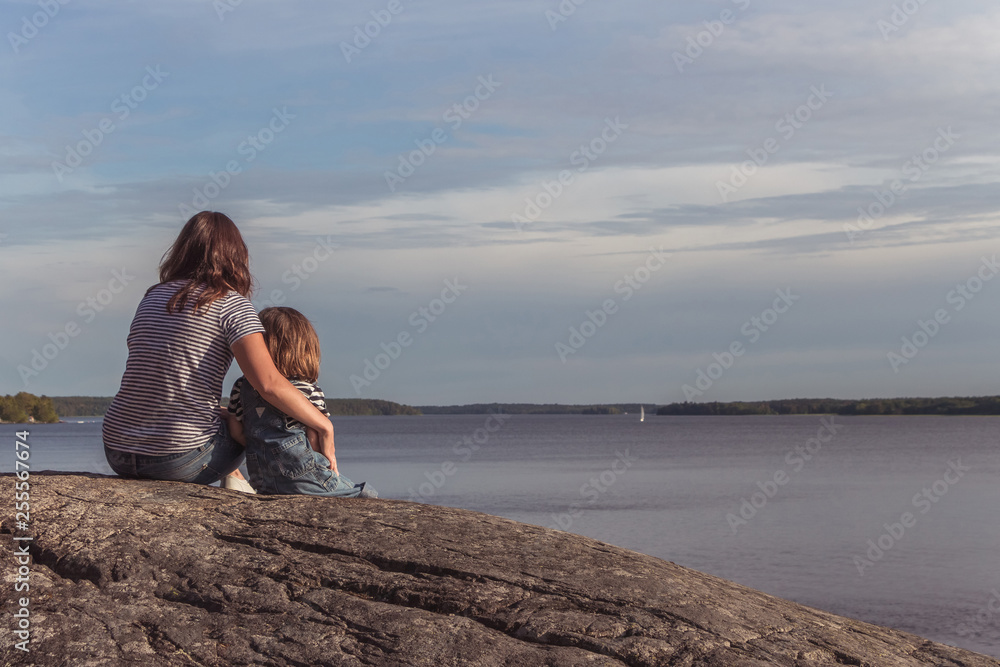 Mom and daughter are sitting and looking at the lake. Mother and little daughter enjoy nature together. Woman and girl by the river. Crown of trees. Blue sky.