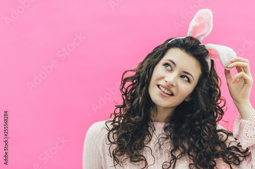 Young pretty girl standing on a pink background. On the head of the ears of the bunny. During this, he holds his ear with his hand and looks up, smiling. Easter.
