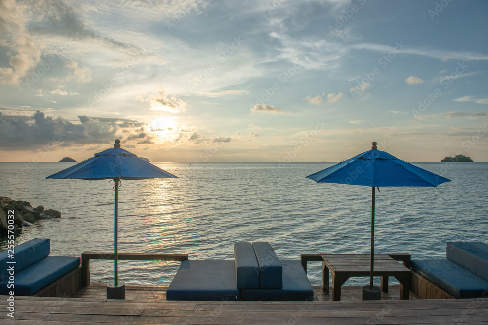 Fototapeta premium Rest on the tropical coast. Umbrellas and two empty lounge chairs stand on a wooden deck near the sea