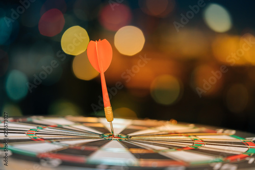 Red dart arrow hitting in the target center of dartboard with city light bokeh background. Target business, achieve and victory concept.
