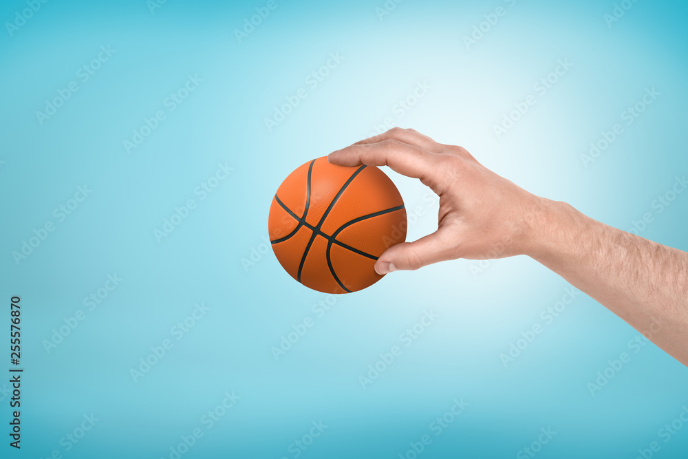 Side close-up of man's hand holding little basketball on light-blue background.