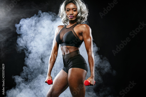 Sporty beautiful black skin woman with dumbbells makes fitness exercising at dark background to stay fit. Sport concept.