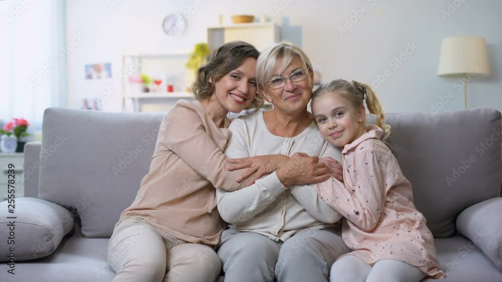 Multi-generation females hugging on home sofa smiling camera, family closeness Multi-generation females hugging on home sofa smiling camera, family closeness