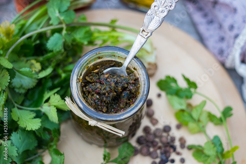 Green adjika. Traditional Georgian spice blend, mix of spices with fresh green coriander, hot chilly pepper in glass jar on wooden board. Food from Georgia.