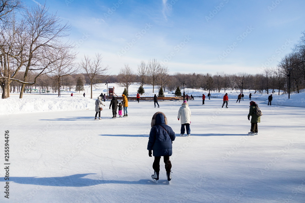Naklejka premium Warmly dressed people skating on frozen lake surrounded by pristine snowy landscape during beautiful sunny day, Beaver Lake, Mount Royal, Montreal, Quebec, Canada