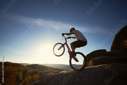 Sportsman cyclist standing on back wheel on trial bicycle, man biker balancing on the top of mountain in the summer sunny morning. Blue sky and sunrise on background. Concept of extreme sport.