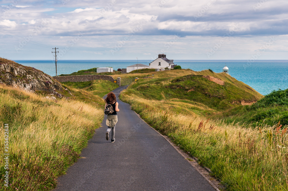 Fototapeta premium Happy young Caucasian female tourist running on an empty road towards the Baily lighthouse on Howth Head, Dublin, Ireland. Irish hills landscape with tall grass and turquoise sea on a summer day.