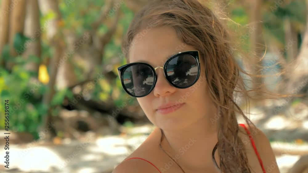 Close-up slow motion portrait of beautiful european young woman in sunglasses, hair blowing in wind on tropical beach, sand and green trees on the background. Travel concept. Close-up slow motion portrait of beautiful european young woman in sunglasses, hair blowing in wind on tropical beach, sand and green trees on the background. Travel concept.
