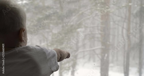 In a slow motion video shoot, a small child (boy) looks out the window on the winter day as snowflakes fall. Concept of: Winter day, Cold day, Mood winter, Snow clapping.