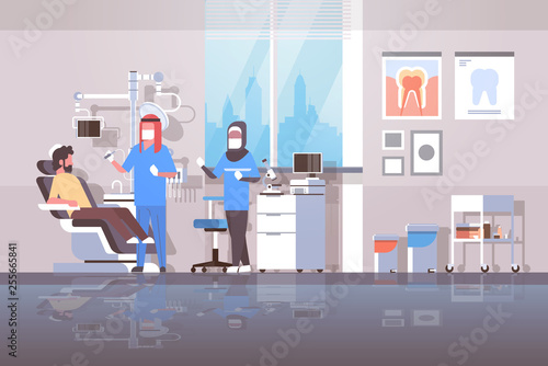 arab dentist with assistant in hijab drilling teeth of man patient lying in dentistry chair professional dental office modern clinic interior arabic characters full length flat horizontal