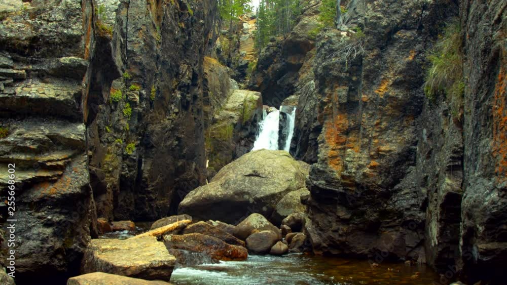 A waterfall cuts through a rocky mountain canyon in summer surrounded by green, lush trees A waterfall cuts through a rocky mountain canyon in summer surrounded by green, lush trees