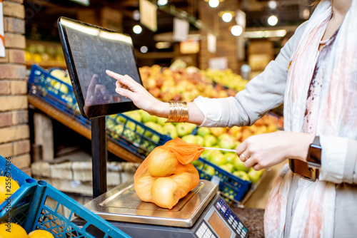 Fototapeta Young woman customer weighing apples packed in eco bag touching the screen of th