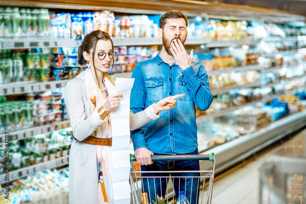 Young couple buying food standing with cart and long shopping list in the supermarket