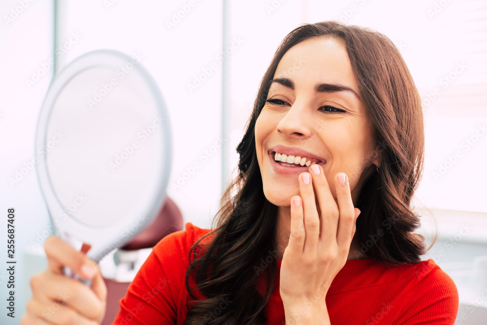 Honed to perfection. Fabulous woman clothed in red sweater, with curly long hair and elegant hands is holding a mirror for looking through the final work of dental doctor. Honed to perfection. Fabulous woman clothed in red sweater, with curly long hair and elegant hands is holding a mirror for looking through the final work of dental doctor.