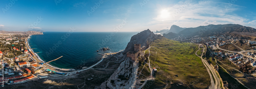 Genoese fortress in Sudak, Crimea. Aerial panorama view of ruins of ancient historic castle on crest of mountain near sea and small town at foot of rocks. Beautiful summer tourist landscape