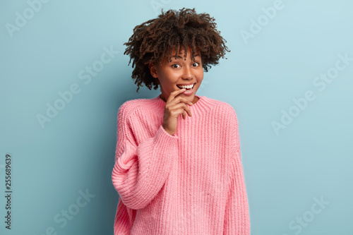 Lovely curious dark skinned female model keeps hands near mouth  looks with happiness and interest  listens intriguing news  dressed in loose light pink sweater  models over blue background.