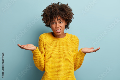 Portrait of unawre unsure dark skinned woman spreads palms in bewilderment, being questioned what to present for parents on holiday, has puzzled facial expression, models over blue background photo
