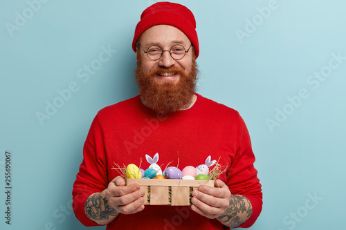 Positive delighted red haired hipster carries small Easter basket with hay and colored eggs, rejoices resurrection and feat Sunday, wears red clothes, round spectacles. Seasonal holiday concept photo