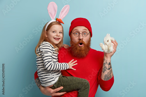 Two family members buy new pet. Surprised ginger bearded dad carries small decorative rabbit with white fur and little smiling daughter, express wonder and happiness, stand against blue background. photo