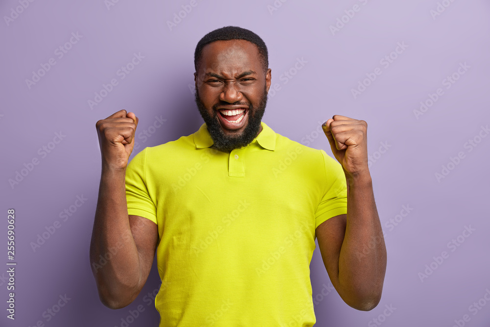 Emotional black ethnic man raises fists with cheer, laughs happily, shows white teeth, shouts for favourite football team, dressed in casual clothes, models over purple background. Winning concept Emotional black ethnic man raises fists with cheer, laughs happily, shows white teeth, shouts for favourite football team, dressed in casual clothes, models over purple background. Winning concept