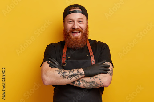 Cheerful male chef in black apron, t shirt and gloves, has thick long ginger beard, keeps arms folded, laughs sincerely, has break after cooking, talks with colleagues in restaurant, shares new recipe photo