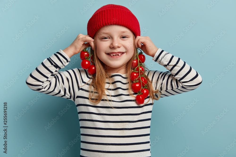 Glad pleasant looking small girl keeps branches of fresh red tomatoes near ears as if earrings, has fun while helps mother to cook dinner, dressed in striped clothes, models indoor. Childhood, eating