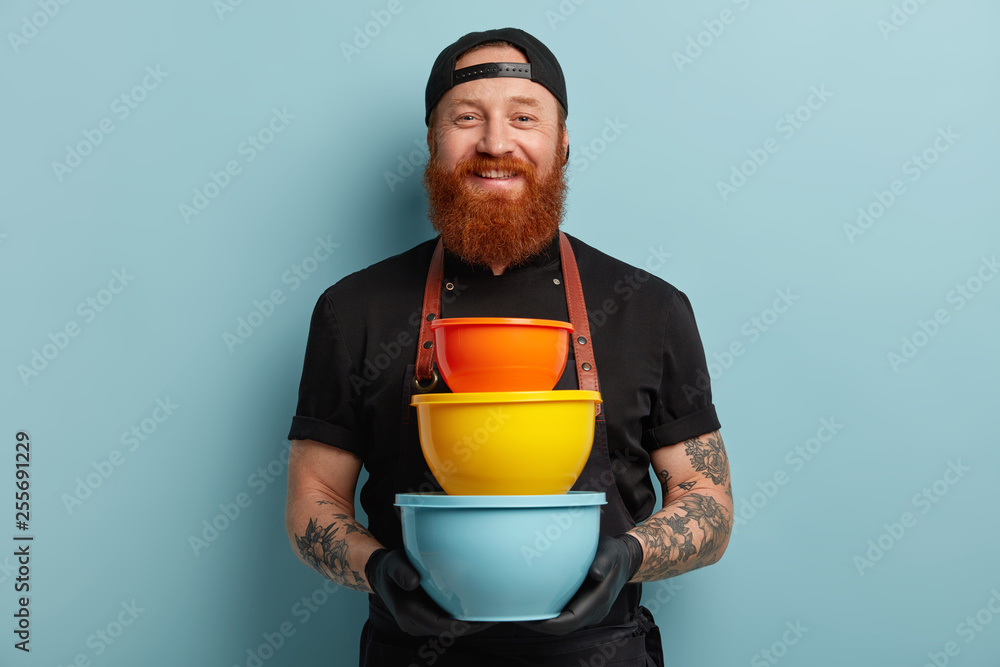 Satisfied male cook holds pile of colorful kitchen utensils, uses dish for cooking dinner, invites you in world of tastes, wears black uniform, rubber protective gloves, isolated on blue background Satisfied male cook holds pile of colorful kitchen utensils, uses dish for cooking dinner, invites you in world of tastes, wears black uniform, rubber protective gloves, isolated on blue background