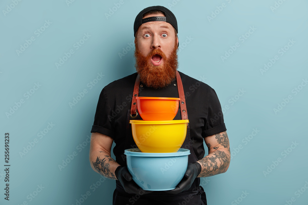 Stupefied emotive tattooed chef holds three plastic dishes, opens mouth widely, has mauch work at kitchen, has red beard, isolated over blue background. Surprised man with colorful pots indoor Stupefied emotive tattooed chef holds three plastic dishes, opens mouth widely, has mauch work at kitchen, has red beard, isolated over blue background. Surprised man with colorful pots indoor