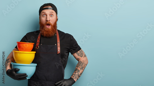 Photo of surprised astonished Caucasian man carries kitchen dish, foregts to buy some products for dinner, keeps hand on waist, dressed in black clothing, keeps jaw dropped from surprisement