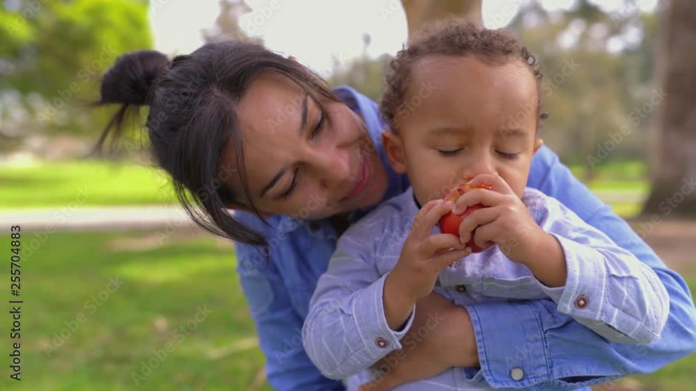 Mixed-race young pretty mother wearing blue jeans and shirt, hugging her little mixed-race son, looking him eating apple. Front view. Family, holiday concept Mixed-race young pretty mother wearing blue jeans and shirt, hugging her little mixed-race son, looking him eating apple. Front view. Family, holiday concept