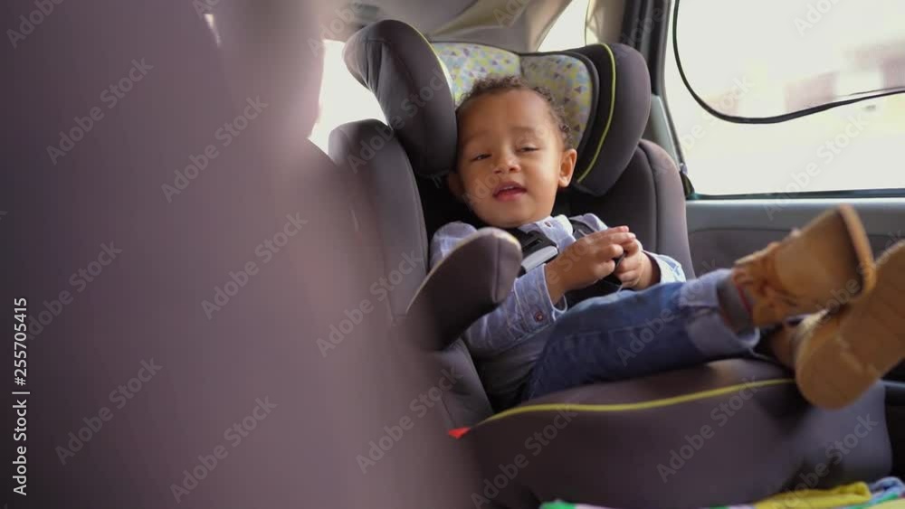 Little mixed-race boy sitting in baby seat, talking to himself, playing with belts. Handsheld shot. Family, safety concept Little mixed-race boy sitting in baby seat, talking to himself, playing with belts. Handsheld shot. Family, safety concept