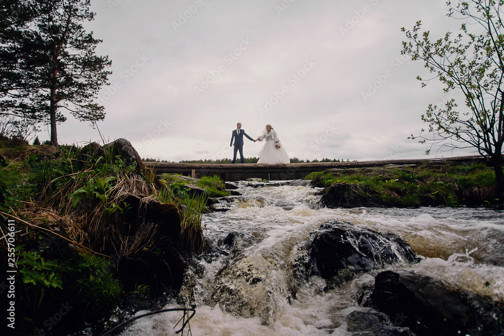 Bride and groom are standing on the bridge over the waterfall. River flows. Couple crossing the river