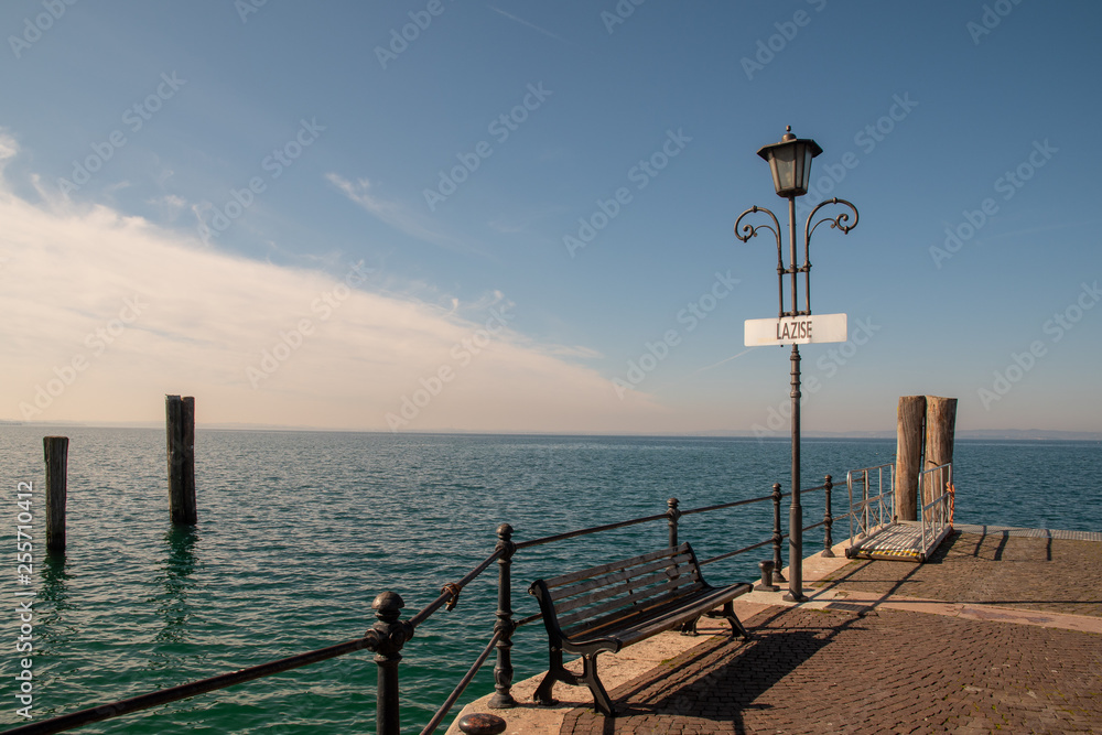 Scenic view of a dock on the shore of Lake Garda with a bench and cloudy blue sky, Lazise, Veneto, Italy Scenic view of a dock on the shore of Lake Garda with a bench and cloudy blue sky, Lazise, Veneto, Italy