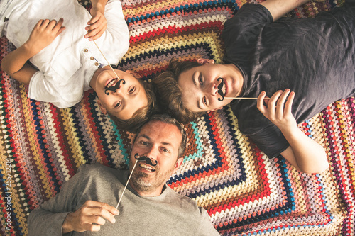 Father and two sons enjoying together lying on a colorful blanket. Tree men of different ages smiling playing with fake mustache. Top view of a couple of teen and their dad. photo