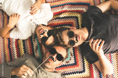 Father and two sons enjoying together lying on a colorful blanket. Tree men of different ages smiling playing with fake mustache. Top view of a couple of teen and their dad with black sunglasses photo