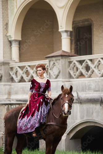 a girl in classical dress pats his horse on the background of the castle