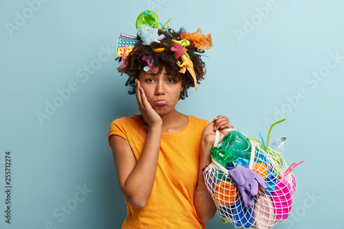 Unhappy dark skinned woman purses lips, has dissatisfied expression, touches cheek, has things made of polymeric material in net bag, plastic products stuck in hair after cleaning. Removing garbage
