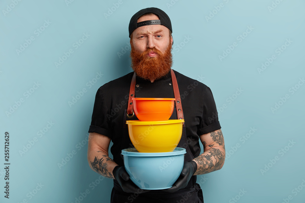 Angry strict male chef holds kitchenware, raises eyebrows, has thick ginger beard, wears cap and apron, buys new containers for kitchen, models over blue background. Cook with colorful dish. Angry strict male chef holds kitchenware, raises eyebrows, has thick ginger beard, wears cap and apron, buys new containers for kitchen, models over blue background. Cook with colorful dish.