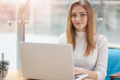 Pretty female student with cute smile prepares for test in cafe. Beautiful happy woman works on laptop computer during coffee break in cafe bar. Blonde wears white sweter sits on blue sofa at window. photo