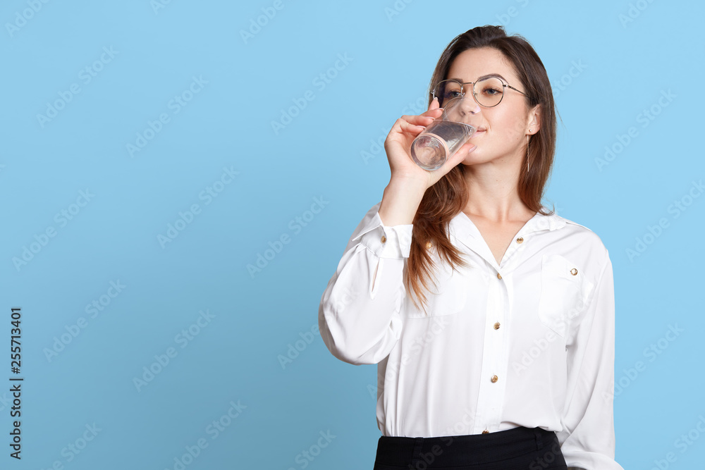 Face portrait of young attractive woman dresses white blouse and black skirt, holds glass in hand and drinks water. Dark haired model wears spectacles poses in studio isolated over blue background. Face portrait of young attractive woman dresses white blouse and black skirt, holds glass in hand and drinks water. Dark haired model wears spectacles poses in studio isolated over blue background.